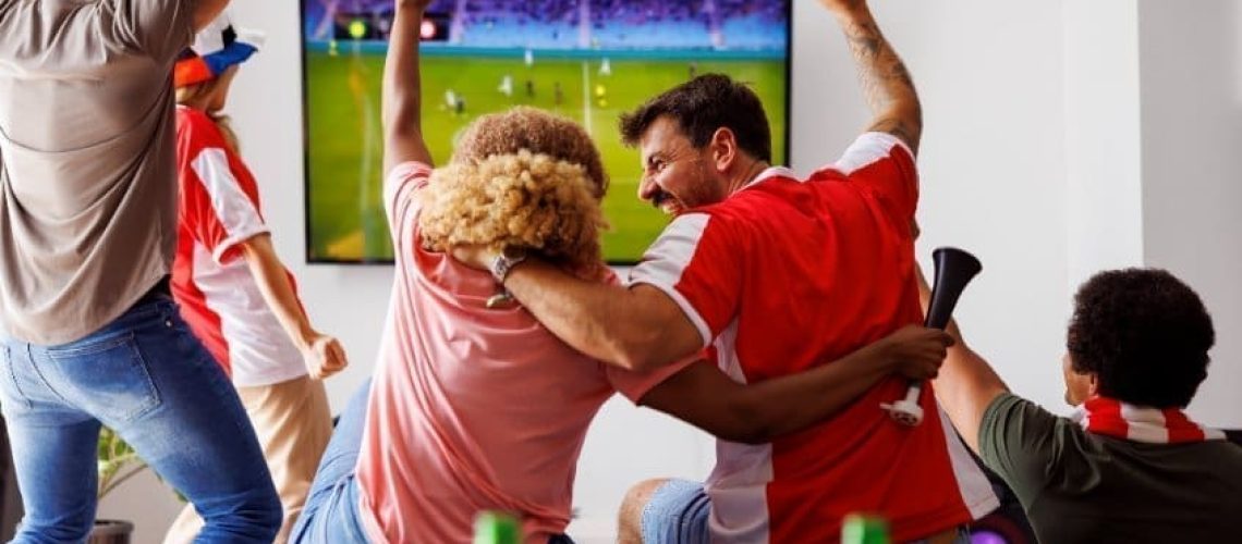 Group of young friends having fun watching football match on TV, drinking beer and cheering; football fans watching game at home celebrating after their team scoring a goal